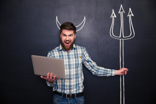 Angry man using laptop and holding trident drawn on blackboard