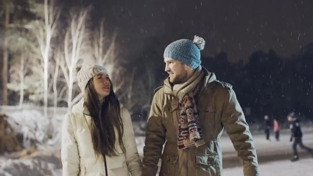Tilt up of romantic young couple skating at outdoor rink holding hands in snowy winter evening 