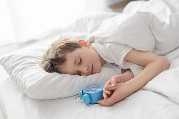 Obraz na płótnie Canvas Little boy sleping in white bed with alarm clock near his head