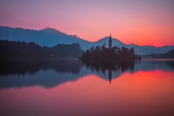 Fototapeta na wymiar Little Island with Catholic Church in Bled Lake, Slovenia at Beautiful Red Sunrise with Castle and Mountains in Background
