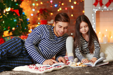 Fototapeta premium Older brother with little sister reading books and eating cookies in Christmas living room