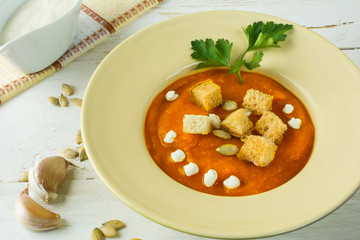 Pumpkin squash vegetable soup with cream, pumpkin seeds, garlic croutons and parsley in a light yellow plate on white wooden background, close up