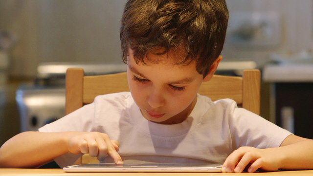 PORTRAIT: A cute little child uses a tablet PC at a table at home. Clothes: white shirt
