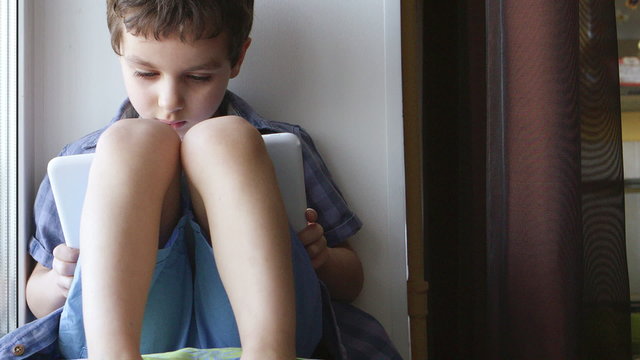 DOLLY: A cute little boy uses a white tablet PC on a windowsill at home
