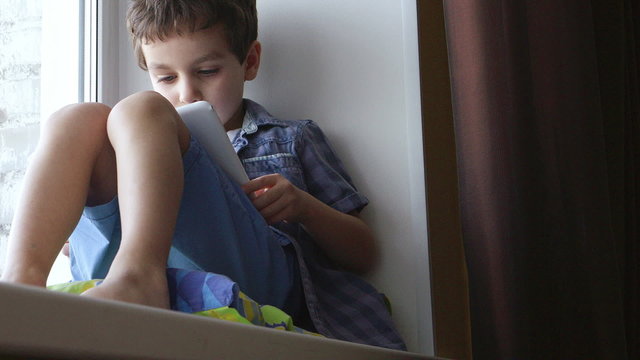 BOTTOM VIEW: A cute little boy uses a white tablet PC on a windowsill at home

