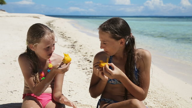 Two girl sisters sitting on a tropical beach,eat mango.children eats mango on the shore of tropical sea on a white beach.Travel concept.Family,summer vacation.Happy family