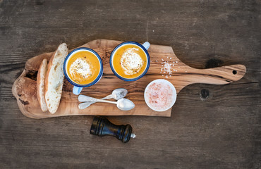 Homemade pumpkin cream soup in enamel mugs with herbs and fresh bread slices on olive serving board over rustic wooden background, horizontal