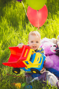 Adorable toddler boy playing toy truck, purple teddy bear and red and green air balloons outside sitting on picnic blanket. Smiling face of happy little boy enjoying nature on sunny spring day.