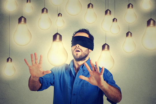 Blindfolded young man walking through light bulbs searching for bright idea