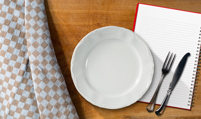 Notebook Plate and Cutlery / Empty plate with silver cutlery and open notebook for recipes or food menu on a wooden table with a brown and white checkered tablecloth
