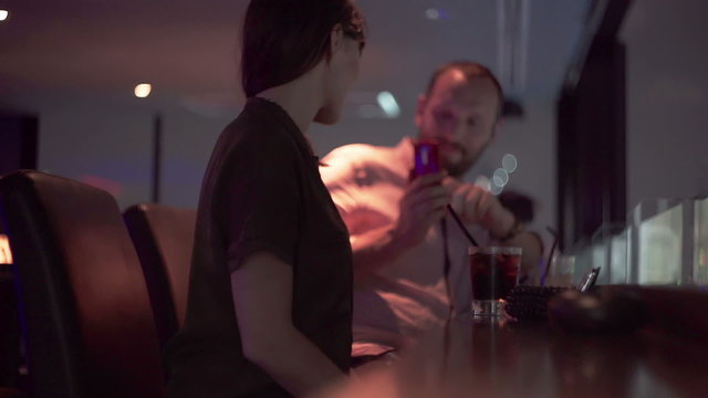 Man taking photo of his beautiful girlfriend with drink in bar at night  
