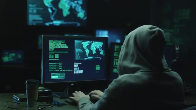 Male hacker in a hood works on a computer with maps and data on display screens in a dark office room. Shot on RED Cinema Camera.