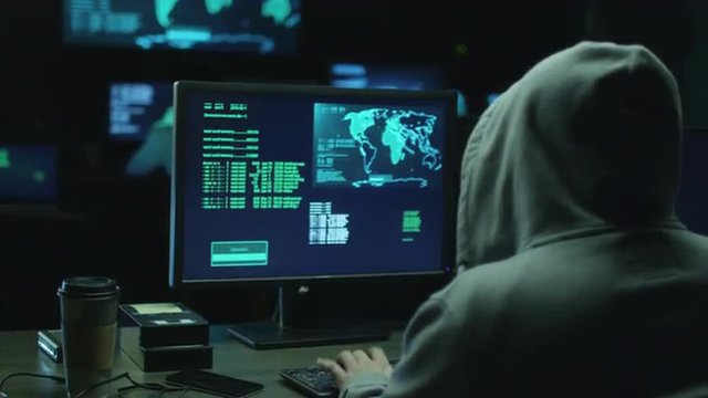 Male hacker in a hood works on a computer with maps and data on display screens in a dark office room. Shot on RED Cinema Camera.
