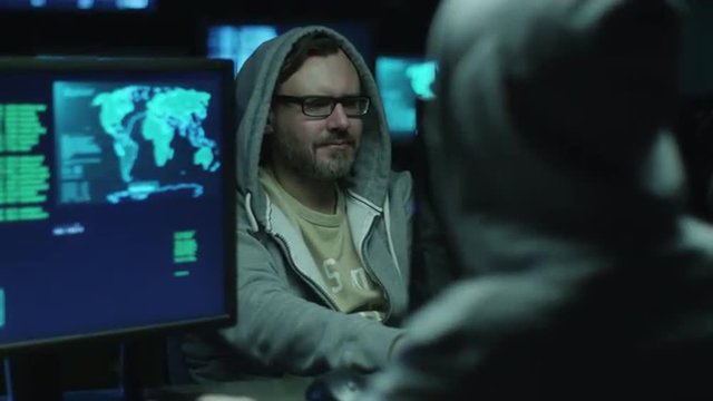 Two hackers in hoods work on a computers with maps and data on display screens in a dark office room. Shot on RED Cinema Camera.