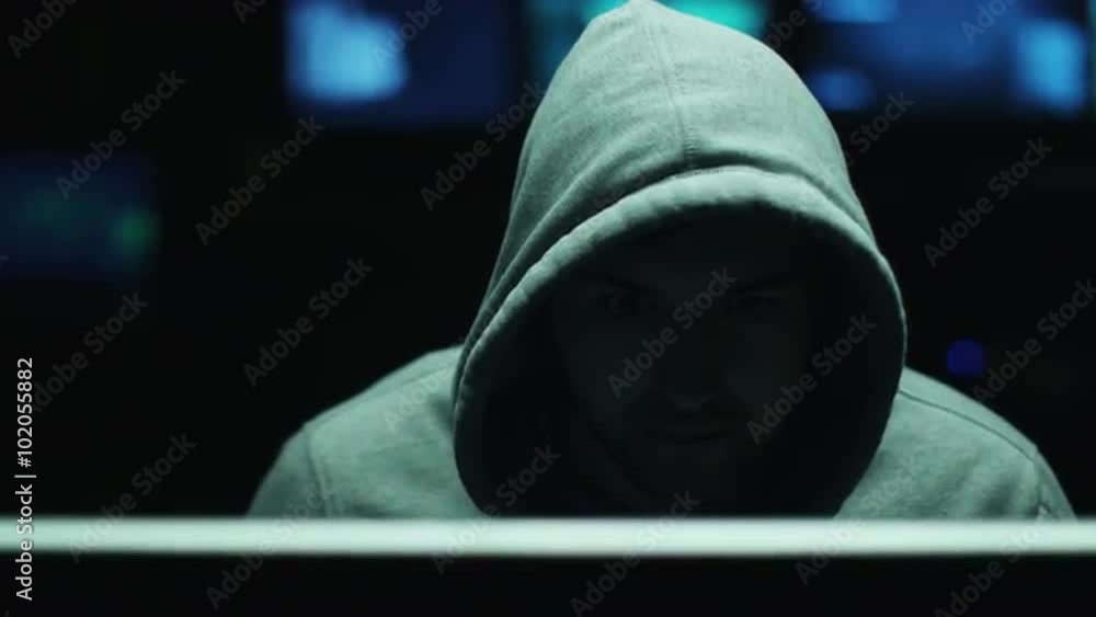 Poster male hacker working on a computer while wearing a hood so his face is hidden from light in a dark of