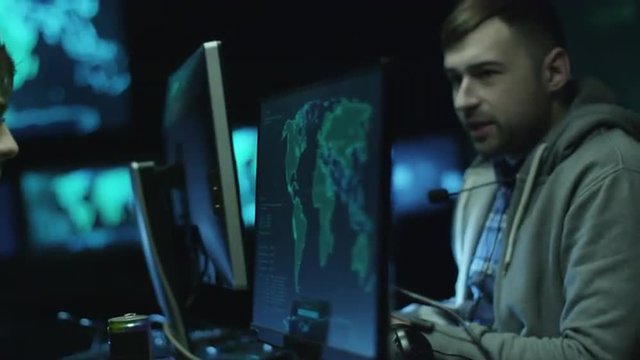 Two hackers in hoodies work on a computers with maps and data on display screens in a dark office room. Shot on RED Cinema Camera.