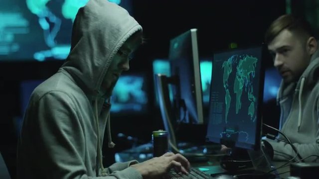 Two hackers in hoodies work on a computers with maps and data on display screens in a dark office room. Shot on RED Cinema Camera.