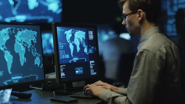 IT professional programmer in glasses is working on computer in cyber security center filled with display screens. Shot on RED Cinema Camera.