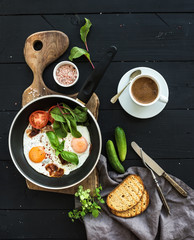 Breakfast set. Pan of fried eggs, bacon, tomatoes with bread, mangold and cucumbers, coffee on rustic wooden serving board over dark table surface