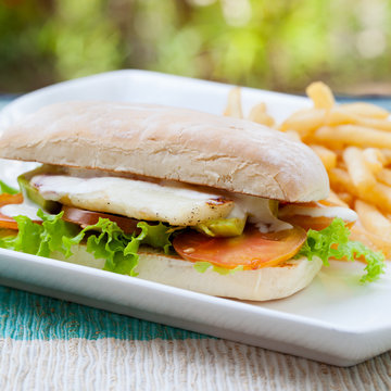 Vegetarian Ciabatta with tomatoes, grilled cheese haloumi, avocado and lettuce with french fries on white plate Summer background