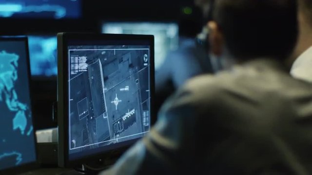 Two IT professionals are spying on businessman through satellite surveillance on computer in a dark monitoring room filled with display screens. Shot on RED Cinema Camera.