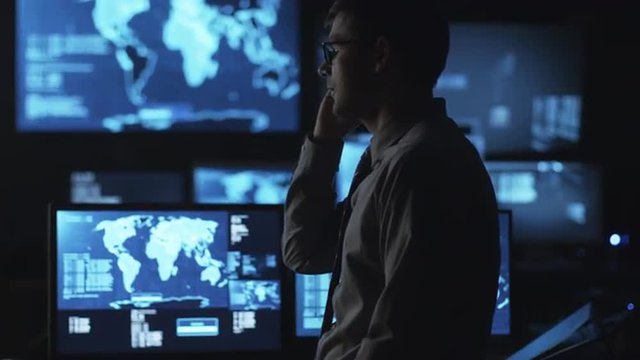 Man In Glasses Is Talking On The Phone In A Dark Monitoring Room Filled With Display Screens. Shot On RED Cinema Camera.