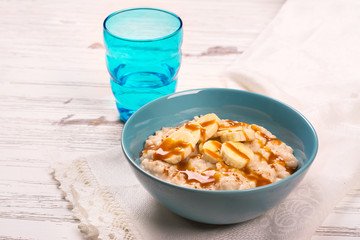 Oatmeal porridge with banana slices, cinnamon and caramel sauce over white wooden table. Selective focus