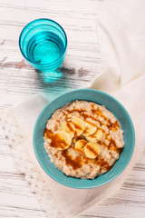 Oatmeal porridge with banana slices, cinnamon and caramel sauce over white wooden table. Selective focus