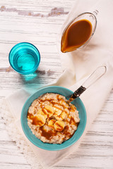 Oatmeal porridge with banana slices, cinnamon and caramel sauce over white wooden table. Selective focus
