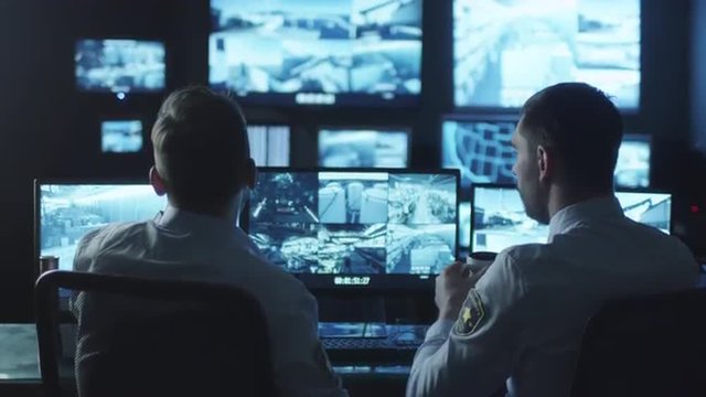 Two security officers are working in a dark monitoring room filled with computer display screens. Shot on RED Cinema Camera.