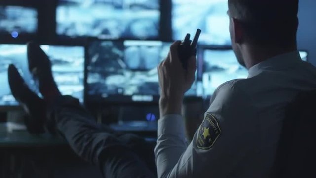 Security officer is relaxing at a computer desk while drinking coffee in a dark monitoring room filled with display screens. Shot on RED Cinema Camera.