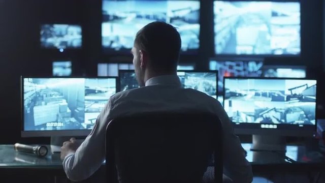 Security officer is drinking coffee while working on a computer in a dark monitoring room filled with display screens. Shot on RED Cinema Camera.