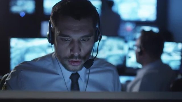 Technical support specialist is talking on a headset while working on a computer in a dark monitoring room filled with display screens. Shot on RED Cinema Camera.