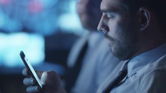 Security officer is checking social media on smartphone while working in a dark monitoring room filled with display screens. Shot on RED Cinema Camera.