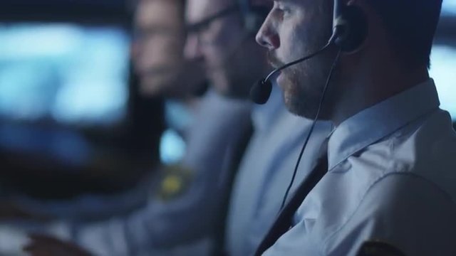 Security officer is talking on a headset while working on a computer in a dark monitoring room filled with display screens. Shot on RED Cinema Camera.