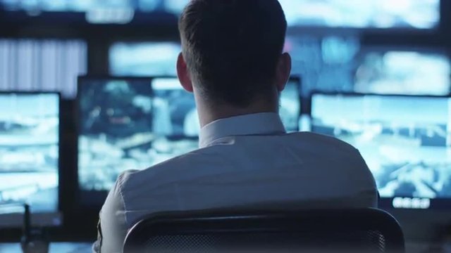 Security officer is working on a computer in a dark monitoring room filled with display screens. Shot on RED Cinema Camera.