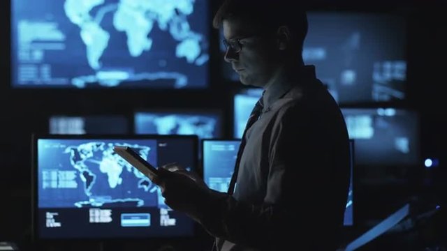 Man in glasses is using a tablet computer in a dark monitoring room filled with display screens. Shot on RED Cinema Camera.