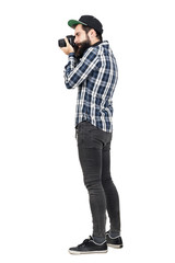 Side view of hipster in plaid shirt and baseball cap taking photo with dslr camera. Full body...