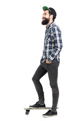 Naklejka premium Side view of bearded hipster with cap standing on skateboard looking away smiling. Full body length portrait isolated over white studio background.