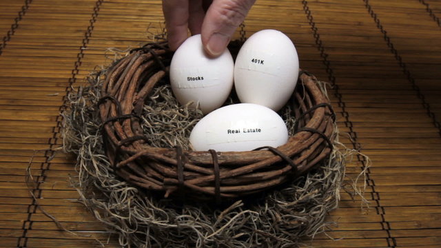 Hand adding egg labeled with stocks to retirement nest with real estate and 401K eggs.
