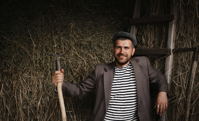 bearded brutal man peasants torn old clothes and a cap with sharp pitchforks throws hay in the barn and drinking alcohol from a bottle © endrews21