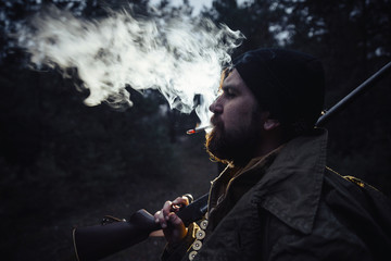 brutal hunter, bearded man in warm hat with a gun in his hand, a knife a backpack and smoking pipe in the dark wild forest in the autumn