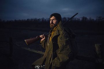 brutal hunter, bearded man in warm hat with a gun in his hand, a knife a backpack and smoking pipe...