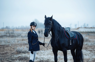 Obraz premium a tall girl with long blond hair in a jockey outfit with a beautiful black horse in an empty snow-covered field in winter