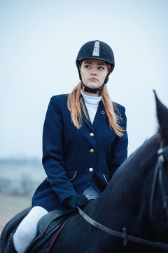 a tall girl with long blond hair in a jockey outfit with a beautiful black horse in an empty snow-covered field in winter