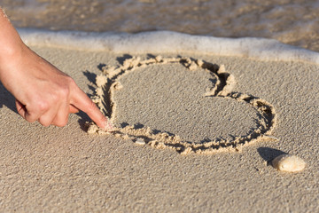 Female hand drawing shape of heart on sandy beach