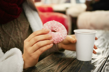 Female hands holding a cup of hot drink and tasty doughnut at the table