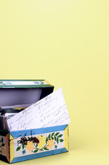 A studio shot of a vintage tin recipe box with a handwritten recipe for Boston Cream Pie showing in the front. A light yellow background with plenty of copy space.