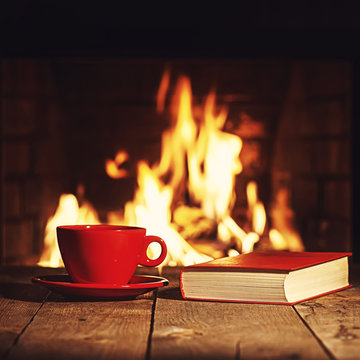 Red cup of coffee or tea and old book on wooden table near  fire