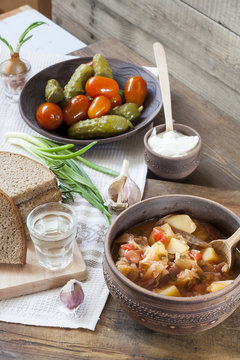 Traditional ukrainian vegetable soup - borsch, marinated tomatoes and cucumbers, sour cream, sliced bread, herbs and garlic, vodka in glass cup at dark wooden table.